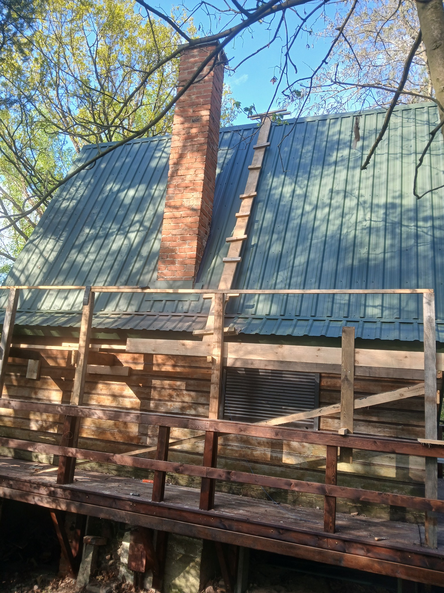 Dom z drewnianą elewacją w trakcie budowy, widoczny komin z cegły i prowizoryczna drabina na dachu. Częściowo wykonana drewniana balustrada.