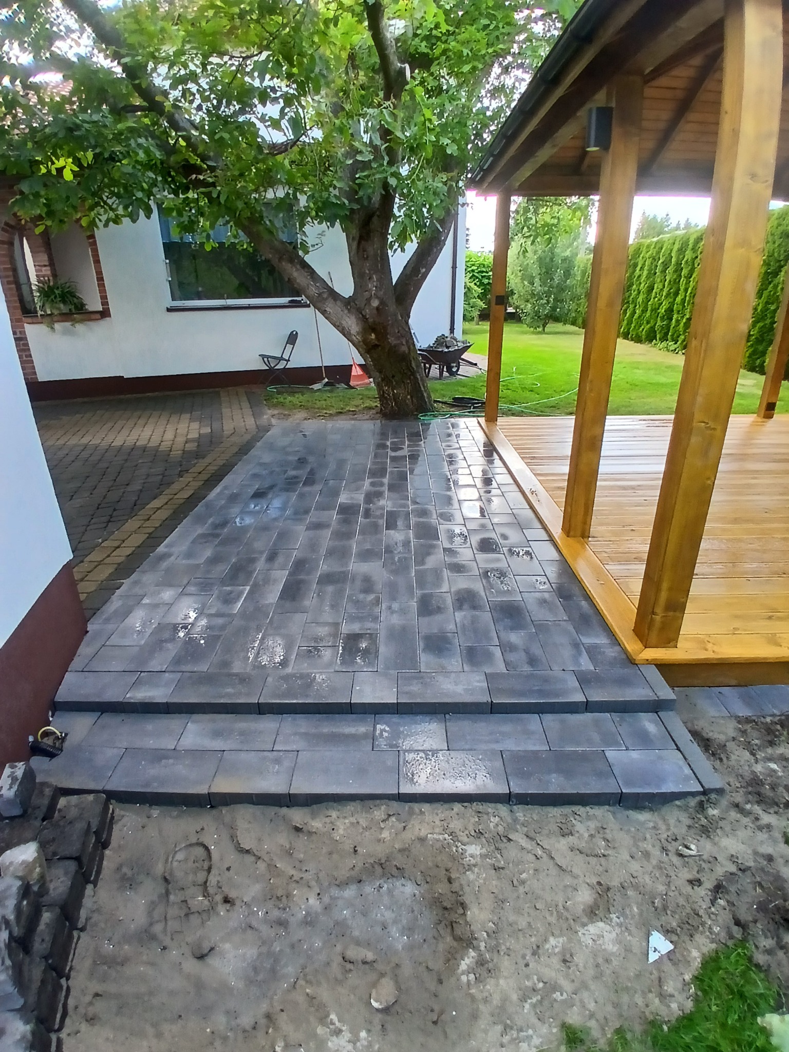 Ułożona kostka brukowa prowadząca do drewnianej pergoli w ogrodzie. Widoczne schody z kostki i fragment domu z oknem. Prace brukarskie w toku.