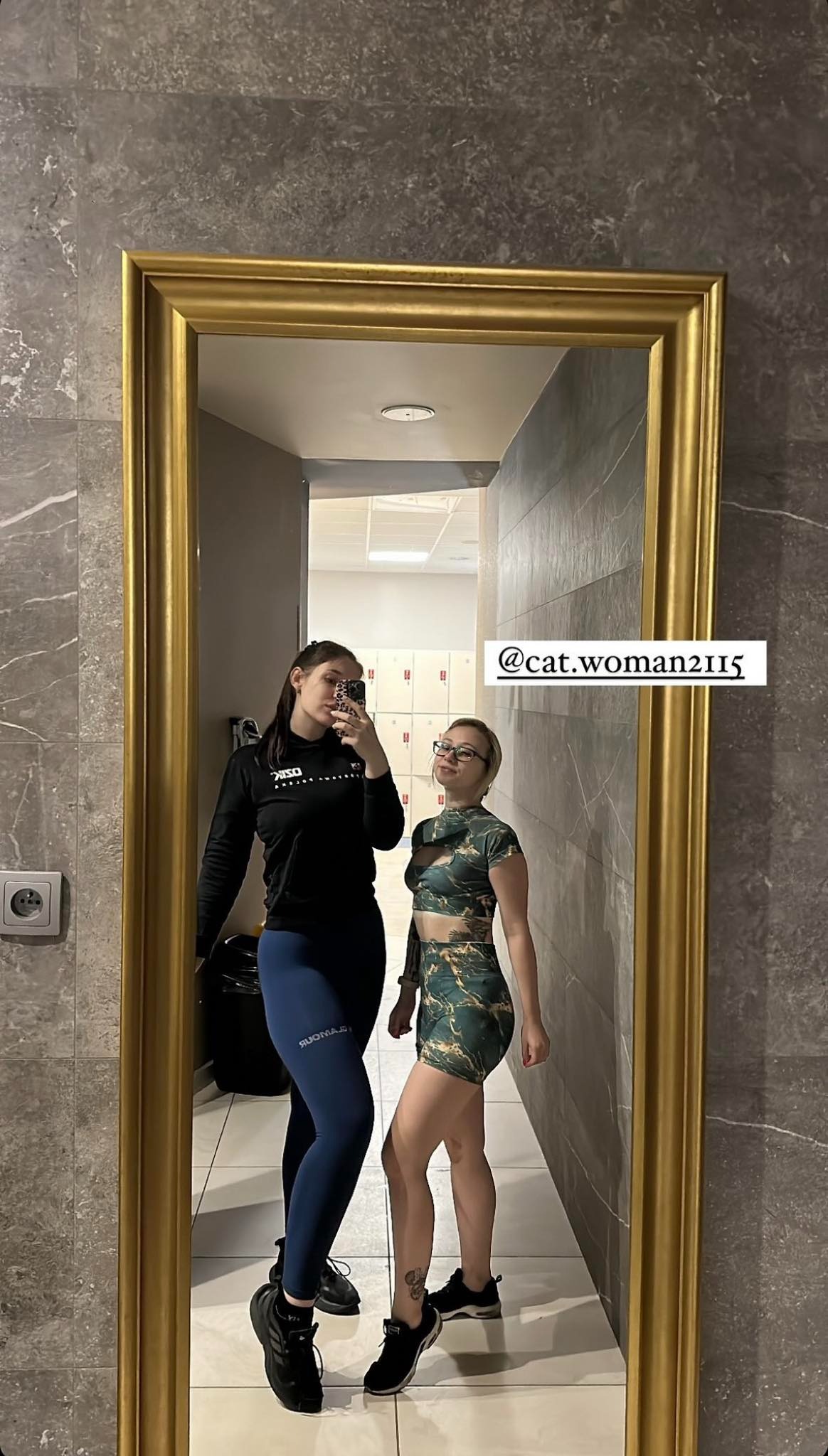 Dwie kobiety w sportowych strojach pozują do zdjęcia w lustrze w szatni z szarymi płytkami i złotą ramą lustra. Jedna w czarnej bluzie i niebieskich legginsach, druga w zielonym komplecie sportowym.