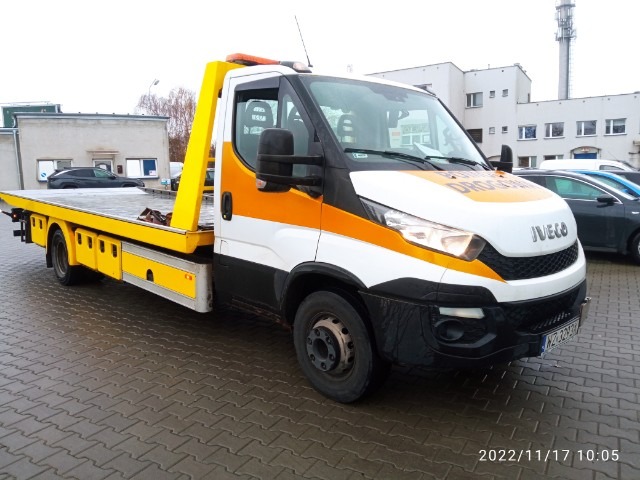 Żółto-biała laweta Iveco z napisem 'Pomoc Drogowa' na masce, zaparkowana na brukowanym placu, widok z boku.