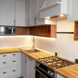 Jasna kuchnia w stylu skandynawskim z drewnianymi blatami i oświetleniem LED podszafkowym, widok na zlew, płytę gazową i piekarnik.