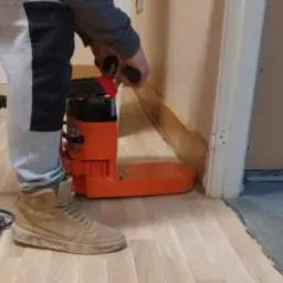 Parkieciarz szlifuje drewnianą podłogę przy użyciu pomarańczowej szlifierki krawędziowej przy ścianie. Widoczny fragment odkurzacza i robocze buty parkieciarza.