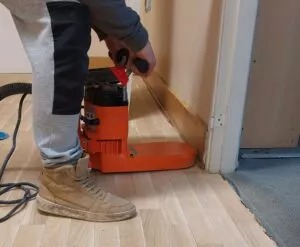 Parkieciarz szlifuje drewnianą podłogę przy użyciu pomarańczowej szlifierki krawędziowej przy ścianie. Widoczny fragment odkurzacza i robocze buty parkieciarza.