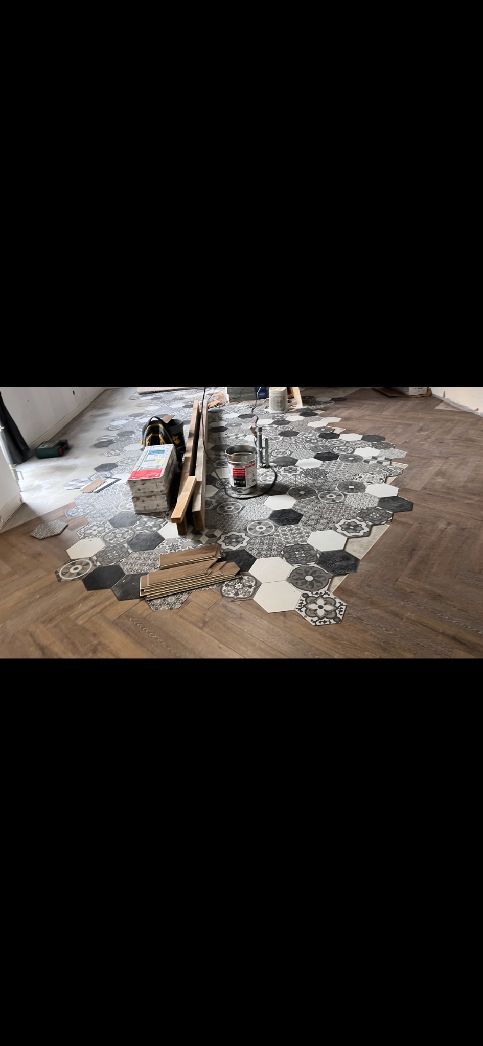 W trakcie układania heksagonalnych płytek podłogowych w odcieniach szarości i bieli na drewnianej podłodze, widoczne narzędzia i materiały montażowe.