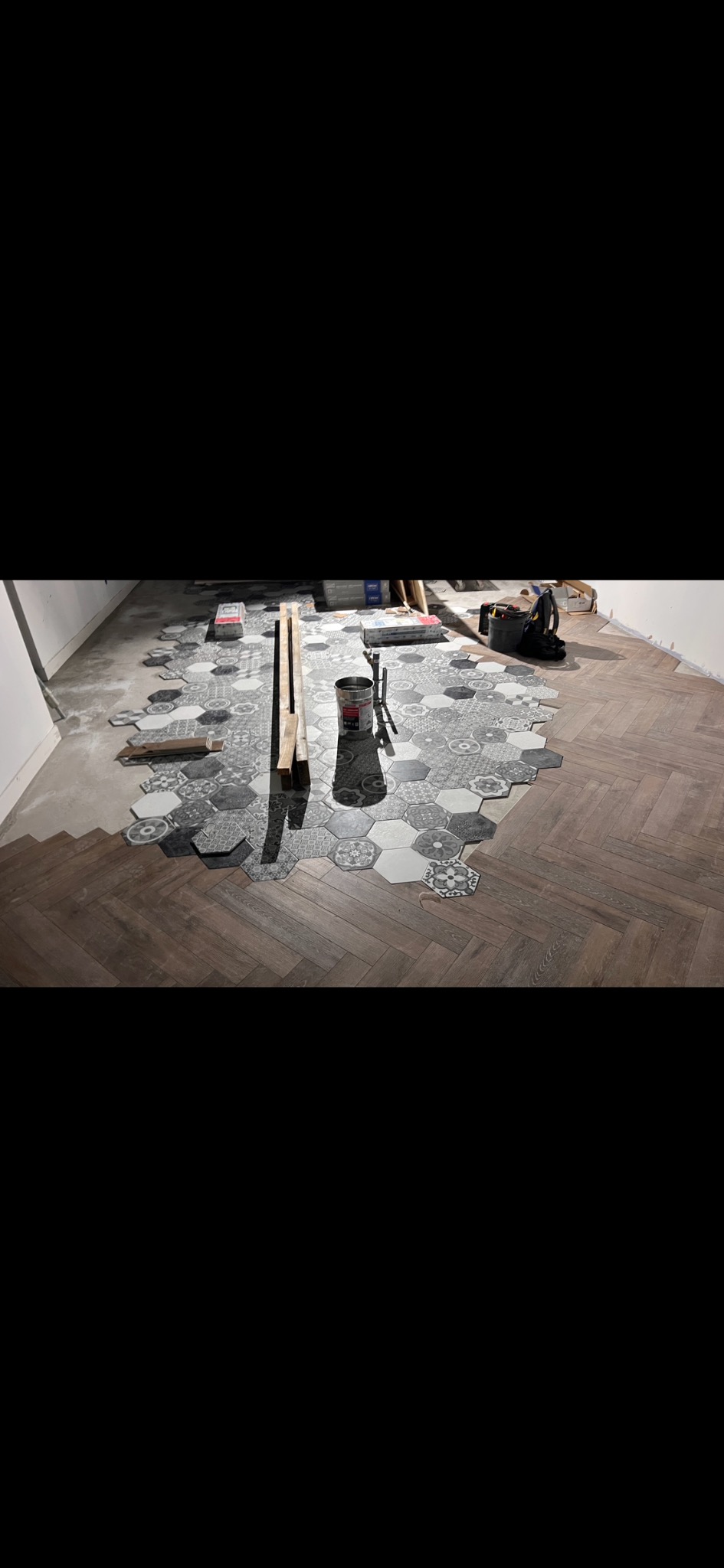 Fragment podłogi z ułożonym parkietem w jodełkę i heksagonalnymi płytkami w odcieniach szarości. Widoczne narzędzia i materiały budowlane. Etap prac wykończeniowych.