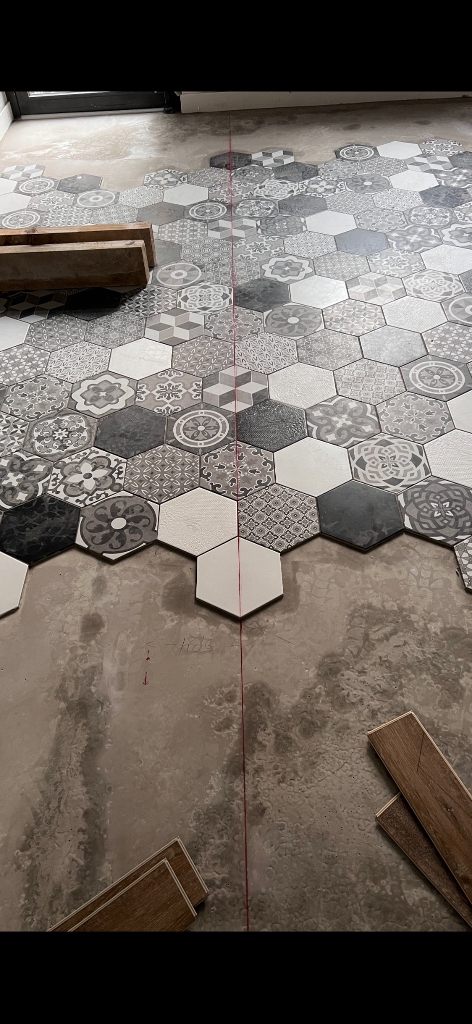 Układanie heksagonalnych płytek podłogowych w odcieniach szarości i bieli, z widoczną czerwoną linią wyznaczającą środek i fragmentami paneli w tle. W trakcie montażu.