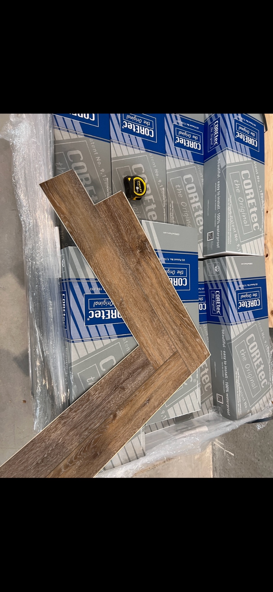 Ułożone panele winylowe w jodełkę na tle zapakowanych paczek paneli Coretec, z widoczną miarką Stanley. Prezentacja wzoru ułożenia paneli podłogowych.