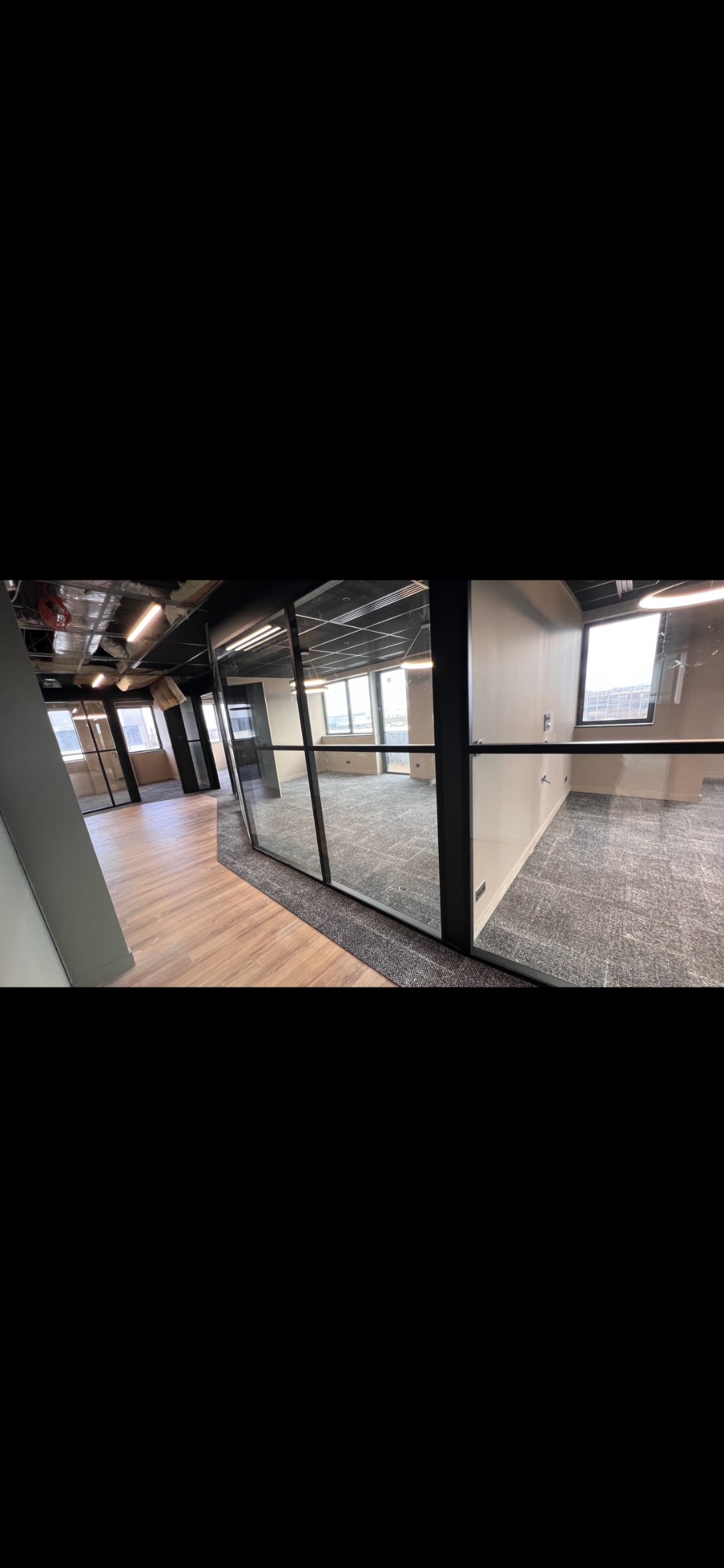 Nowoczesne biuro po remoncie: podłoga z paneli i wykładzina dywanowa oddzielone szklaną ścianą działową. Widok na okna i oświetlenie sufitowe.