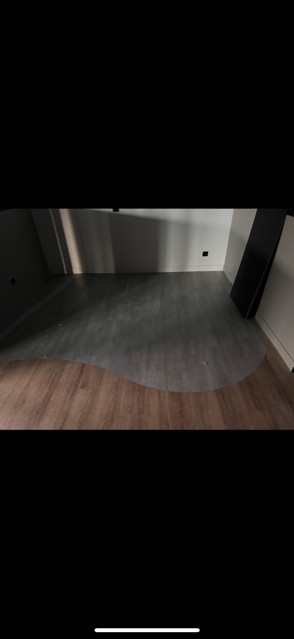 Fragment podłogi z nietypowym połączeniem paneli drewnopodobnych i szarych płytek, tworzącym falistą linię przejścia. Widoczne listwy przypodłogowe i gniazdka.