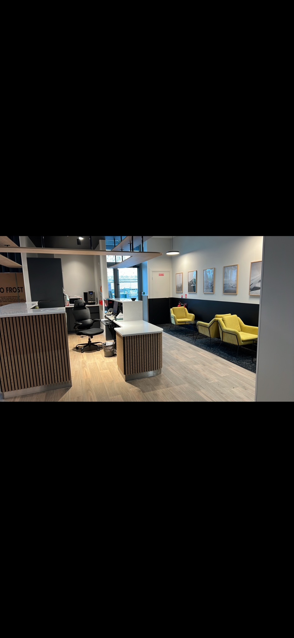 Nowoczesne wnętrze biurowe z drewnianymi panelami na ladzie recepcyjnej i podłodze, żółte fotele w strefie oczekiwania, czarno-białe ściany i grafiki.