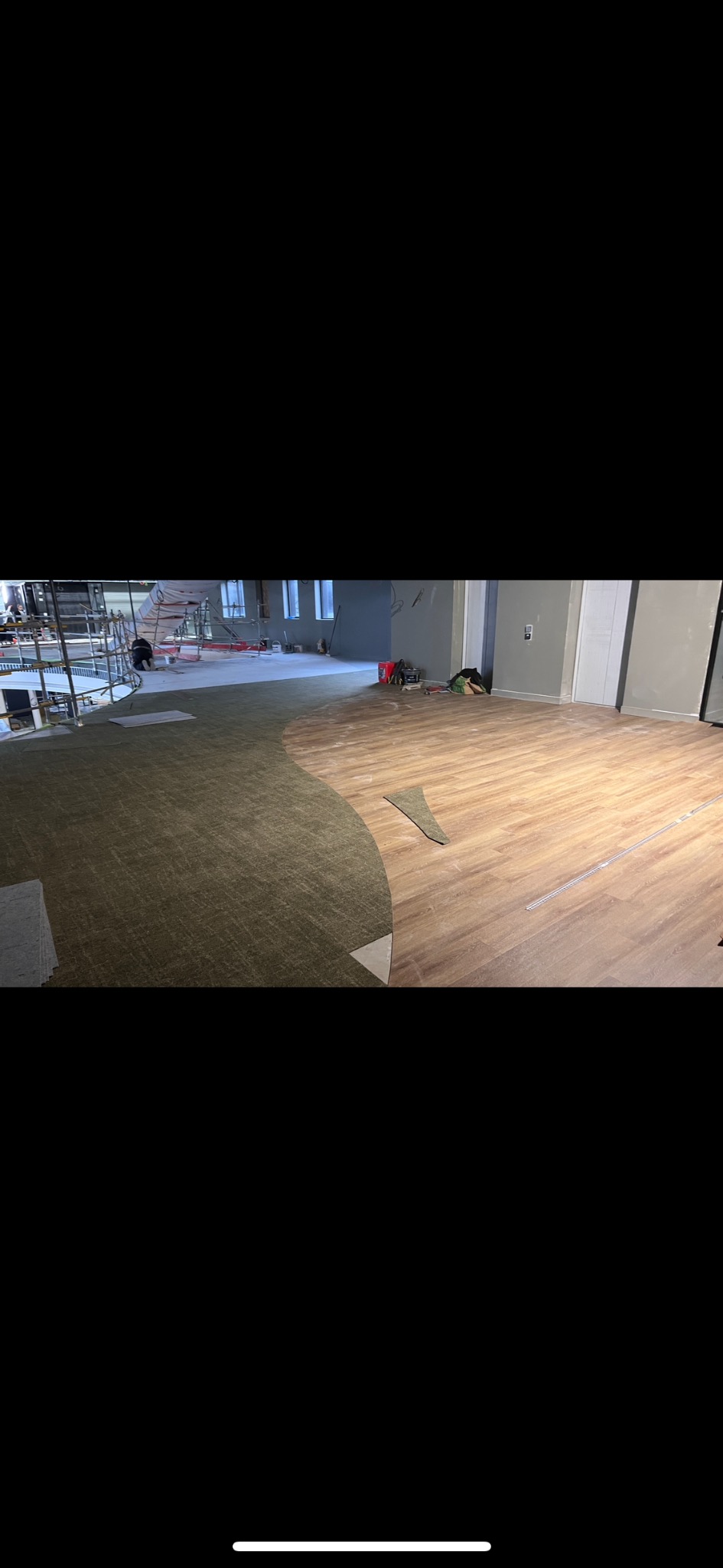 Układanie podłogi: połączenie zielonej wykładziny z panelami drewnopodobnymi w przestronnym wnętrzu. Widoczne narzędzia pracy i fragment rusztowania w tle.