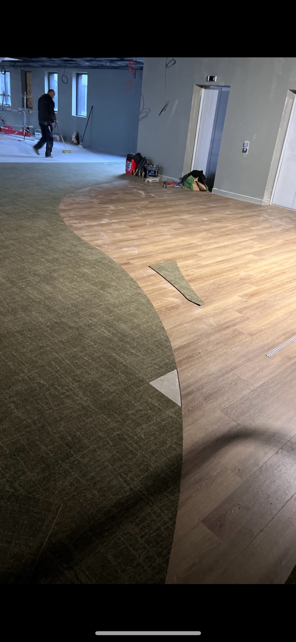 Układanie podłogi: fragment wykładziny dywanowej łączy się z panelami imitującymi drewno. W tle widoczny pracownik i narzędzia.