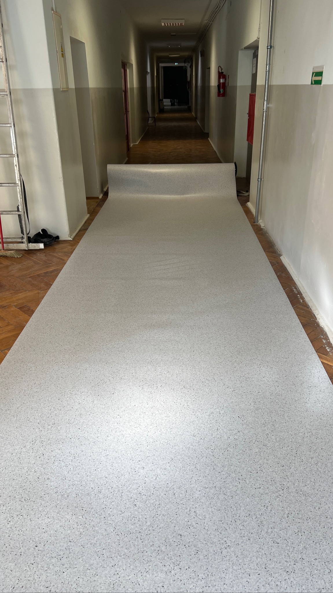 Długi korytarz z częściowo rozłożoną, jasną wykładziną w drobny wzór. Widoczna drabina i gaśnica na ścianie. Podłoga w korytarzu wyłożona parkietem.