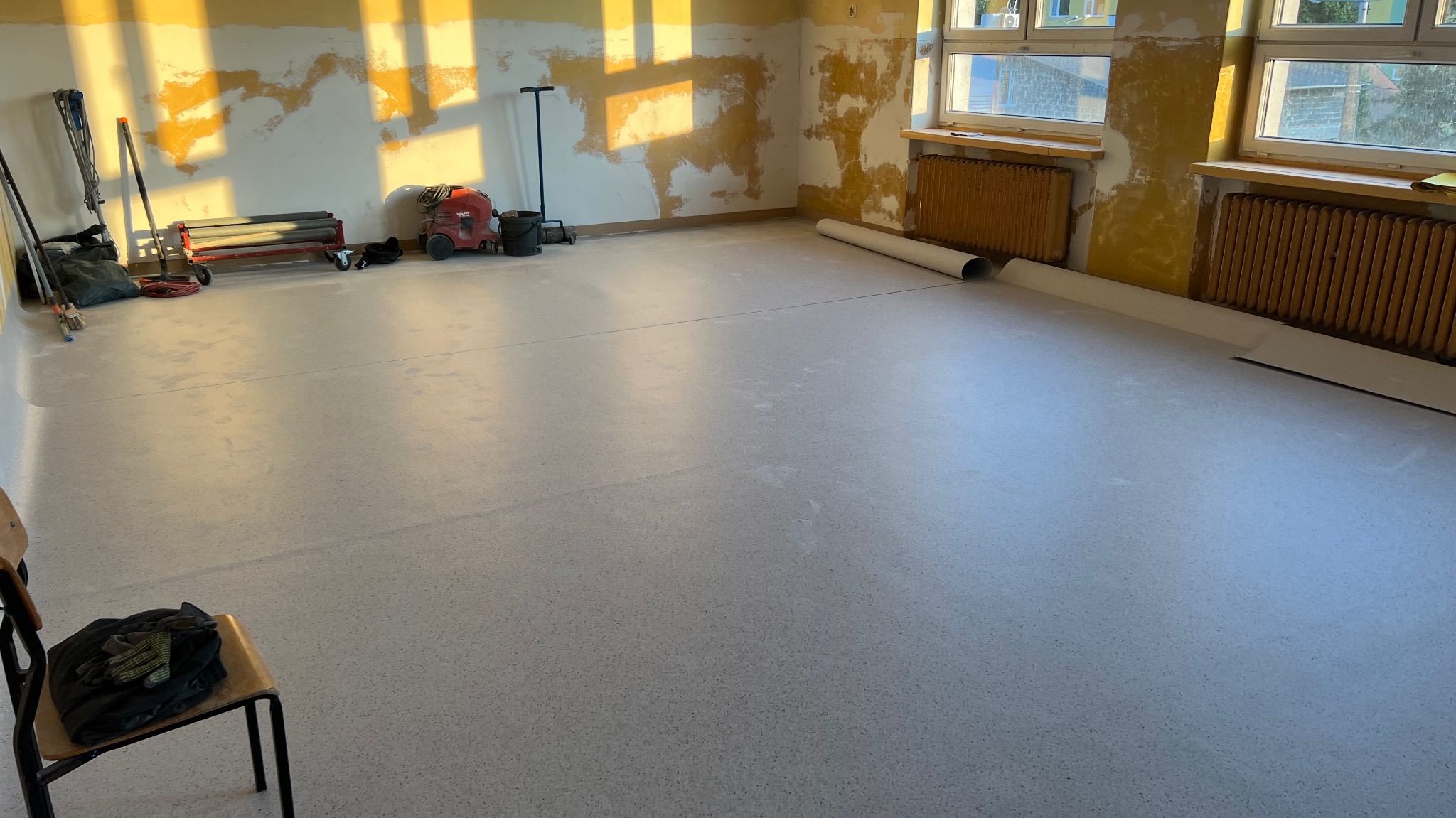 Jasna wykładzina w pokoju z przygotowaną ścianą do malowania. Widoczne narzędzia i wałek wykładziny. Promienie słońca na ścianie.