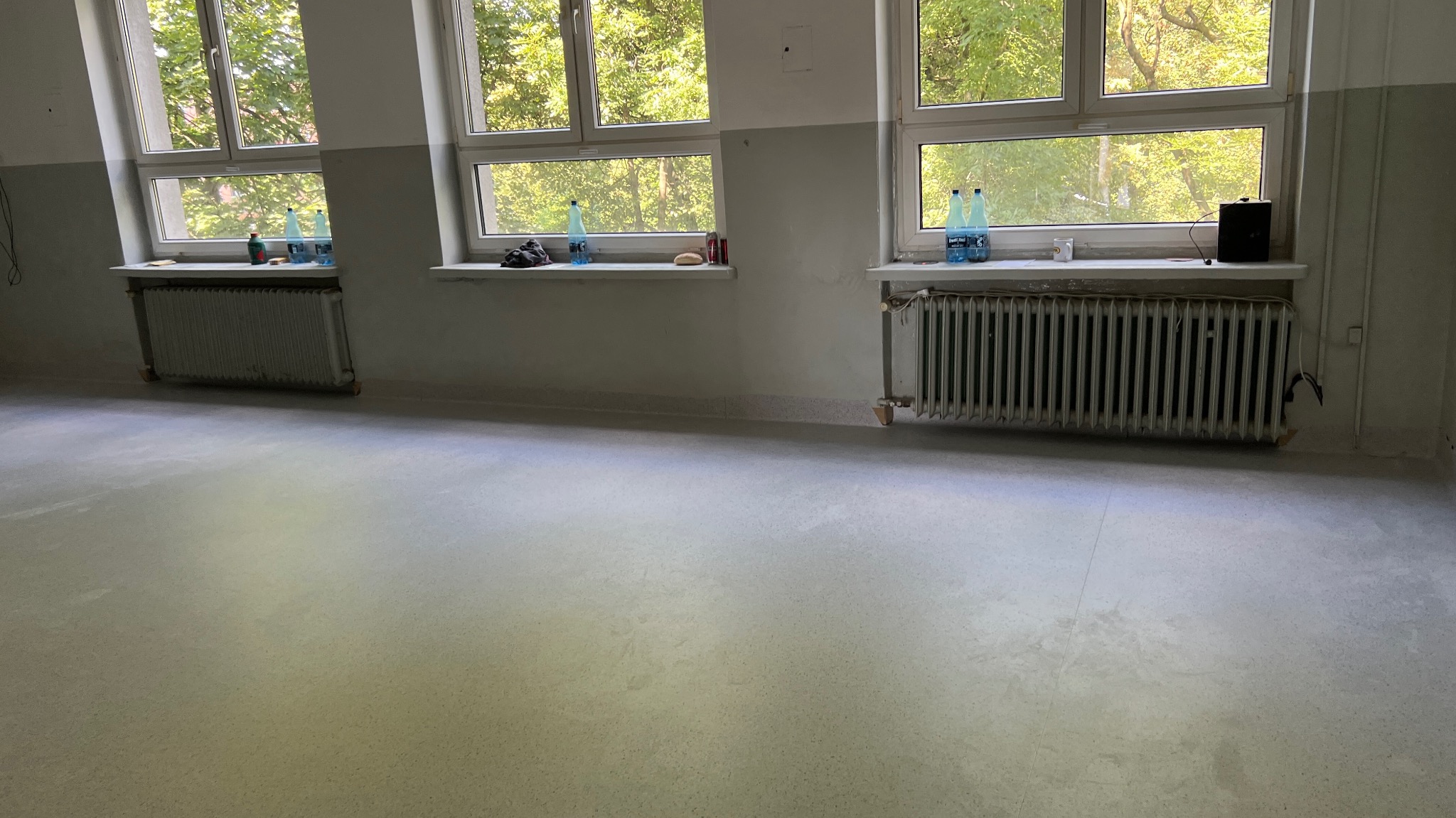 Jasne, puste pomieszczenie po remoncie podłogi, z trzema oknami i grzejnikami. Widoczne butelki z wodą na parapetach. Świeżo położona, jasna wykładzina podłogowa.