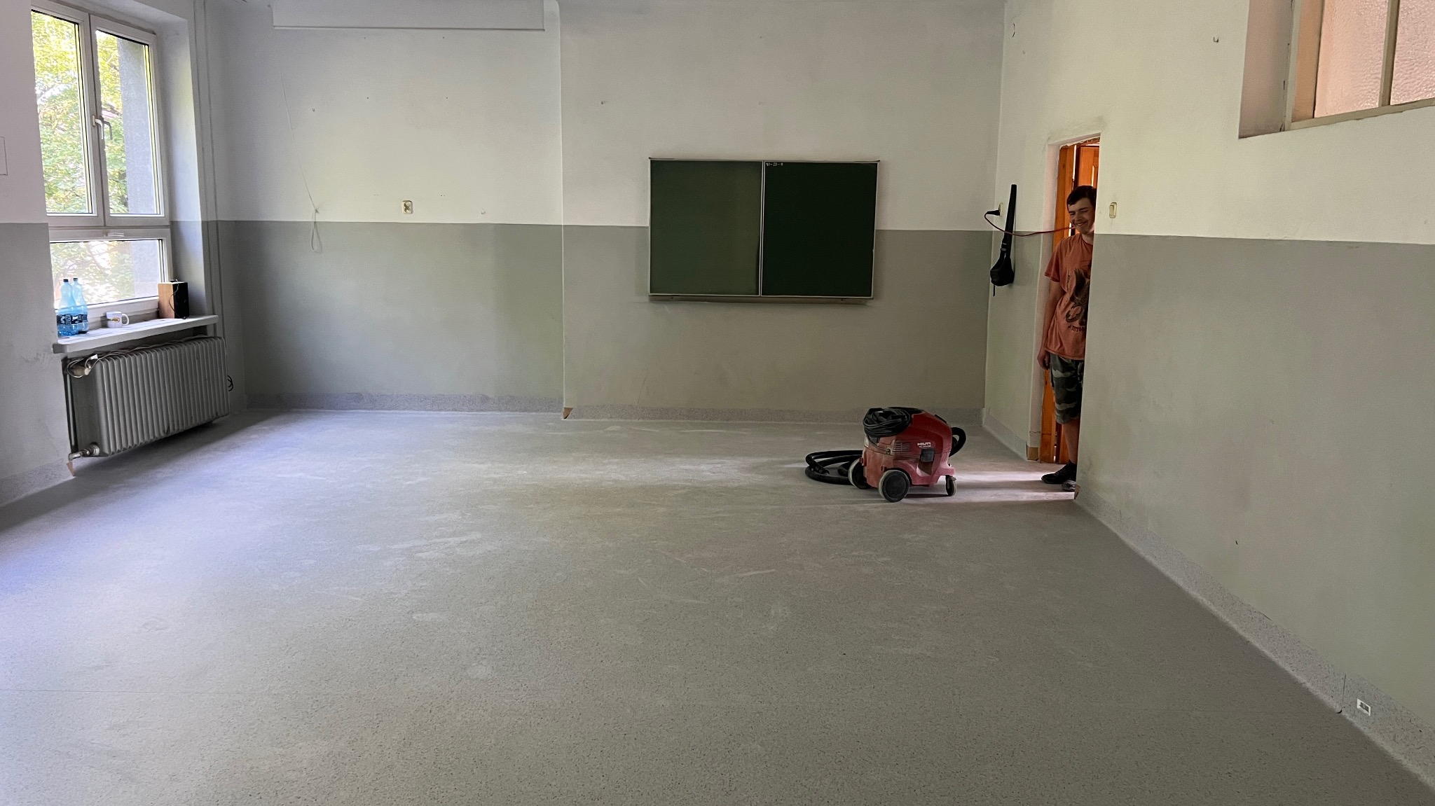 Jasne pomieszczenie po remoncie z nową, szarą podłogą. Widoczny odkurzacz przemysłowy i fragment osoby w drzwiach. Ściany dwukolorowe, okno z lewej strony.