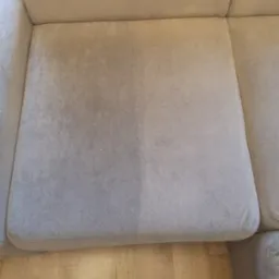 Fragment siedziska sofy z widoczną różnicą w czystości po lewej stronie, ukazując efekt czyszczenia tapicerki, na tle drewnianej podłogi.