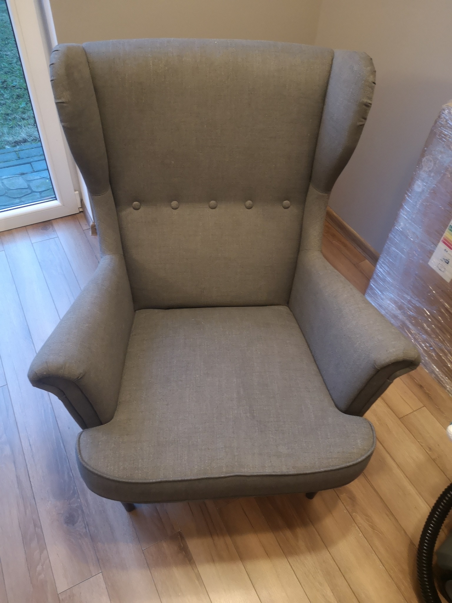 Szary fotel typu uszak z czterema guzikami w oparciu, stojący na drewnianej podłodze, obok okna i zapakowanego przedmiotu.