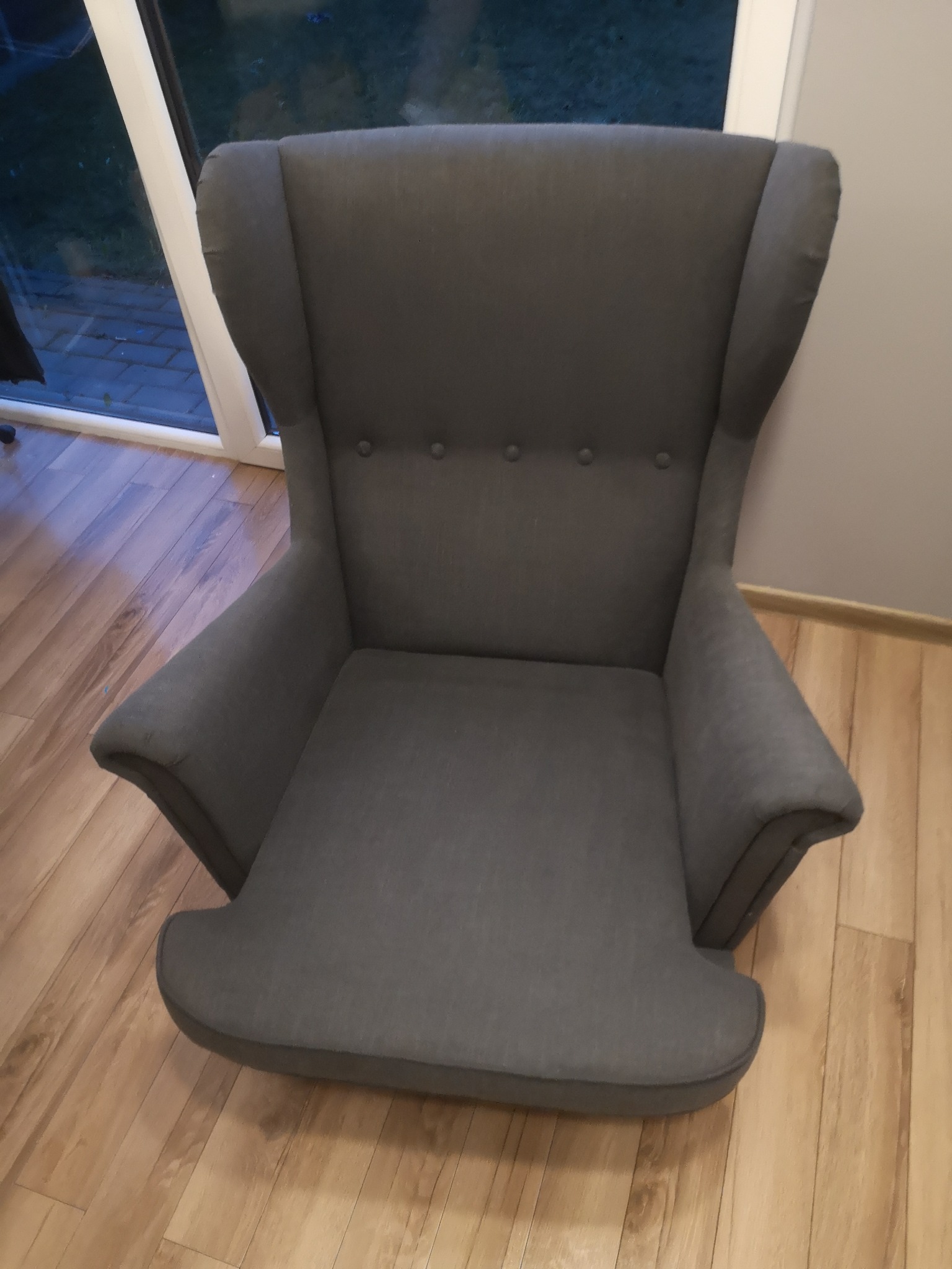 Szary fotel typu uszak z tapicerką, widoczny na tle drewnianej podłogi i fragmentu okna.