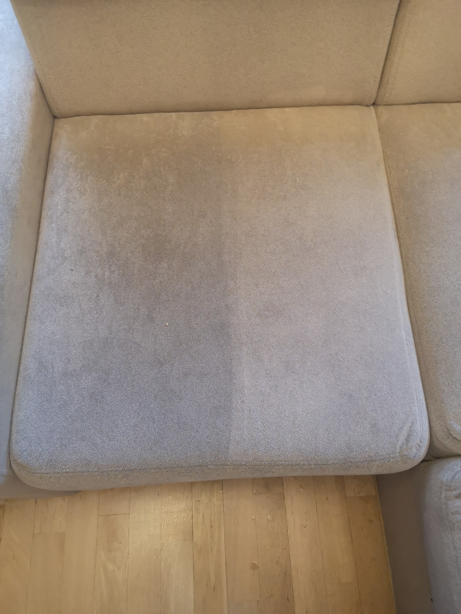 Fragment siedziska sofy z widoczną różnicą w czystości po lewej stronie, ukazując efekt czyszczenia tapicerki, na tle drewnianej podłogi.