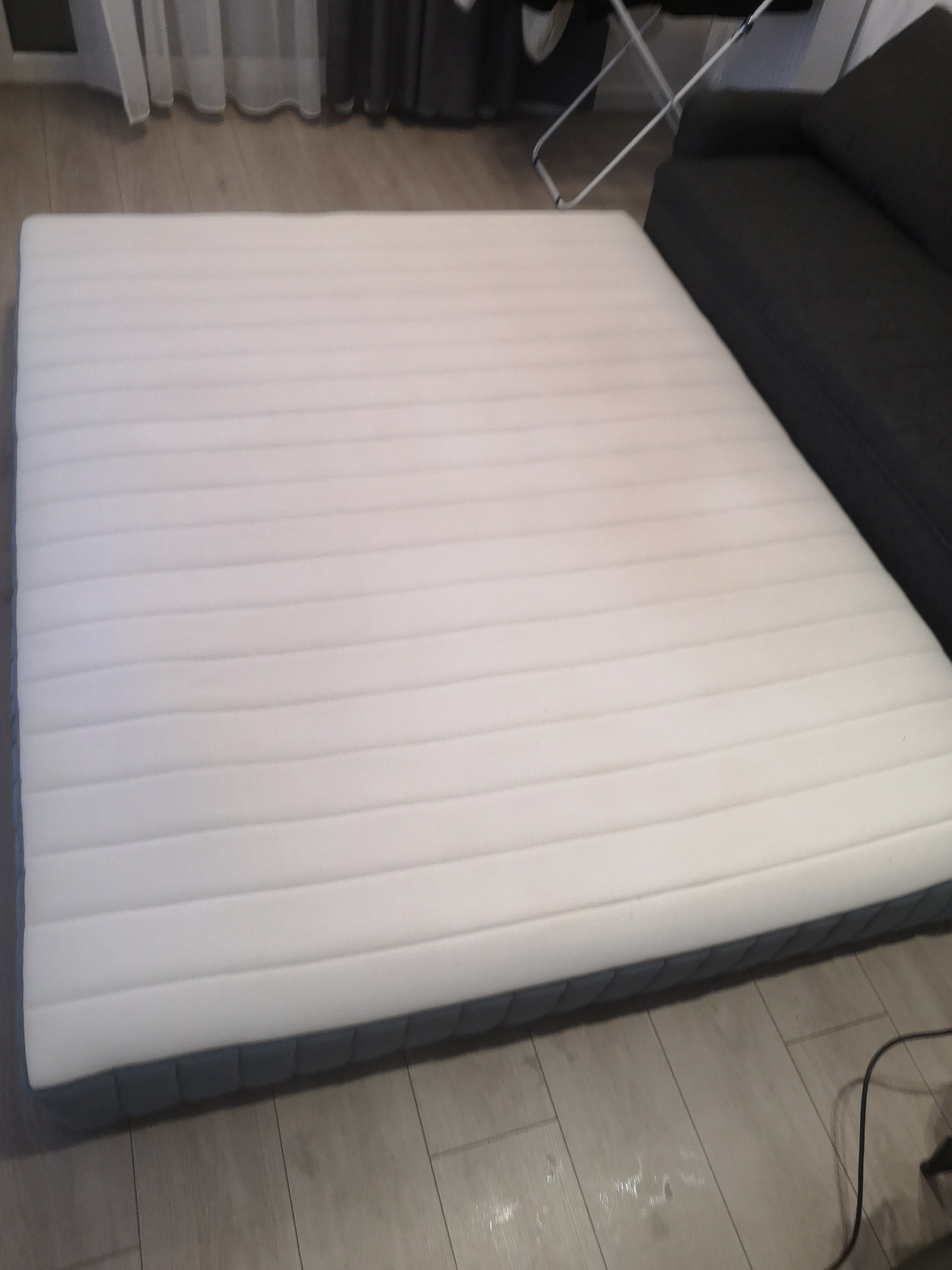 Biały materac z widocznymi śladami użytkowania leżący na podłodze obok szarej sofy.