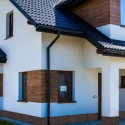 Nowoczesny dom jednorodzinny z białą elewacją, brązowymi panelami dekoracyjnymi i ciemną dachówką, widoczny z zewnątrz w słoneczny dzień.