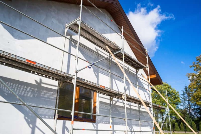 Elewacja domu w trakcie prac, widoczne rusztowanie i częściowo otynkowana ściana w jasnym kolorze, okno z brązową ramą, błękitne niebo w tle.