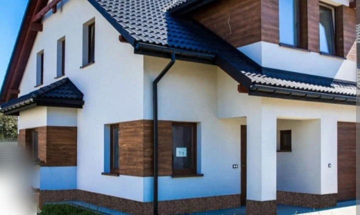 Nowoczesny dom jednorodzinny z białą elewacją, brązowymi panelami dekoracyjnymi i ciemną dachówką, widoczny z zewnątrz w słoneczny dzień.