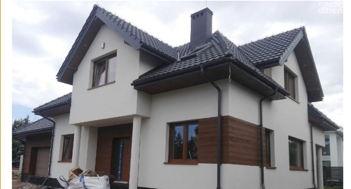 Dwukondygnacyjny dom jednorodzinny z jasną elewacją i fragmentami okładziny drewnopodobnej, ciemna dachówka i orynnowanie, widoczne worki z materiałami budowlanymi.