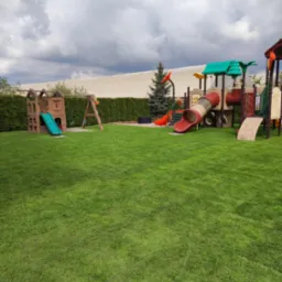 Rozległy, zielony trawnik z równo skoszoną trawą, otoczony żywopłotem i wyposażony w kolorowy plac zabaw z drewnianą konstrukcją, zjeżdżalniami i huśtawkami, na tle pochmurnego nieba.