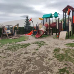 Żółta koparka pracuje na placu zabaw, przygotowując teren pod instalację urządzeń, w tle widoczny duży namiot.