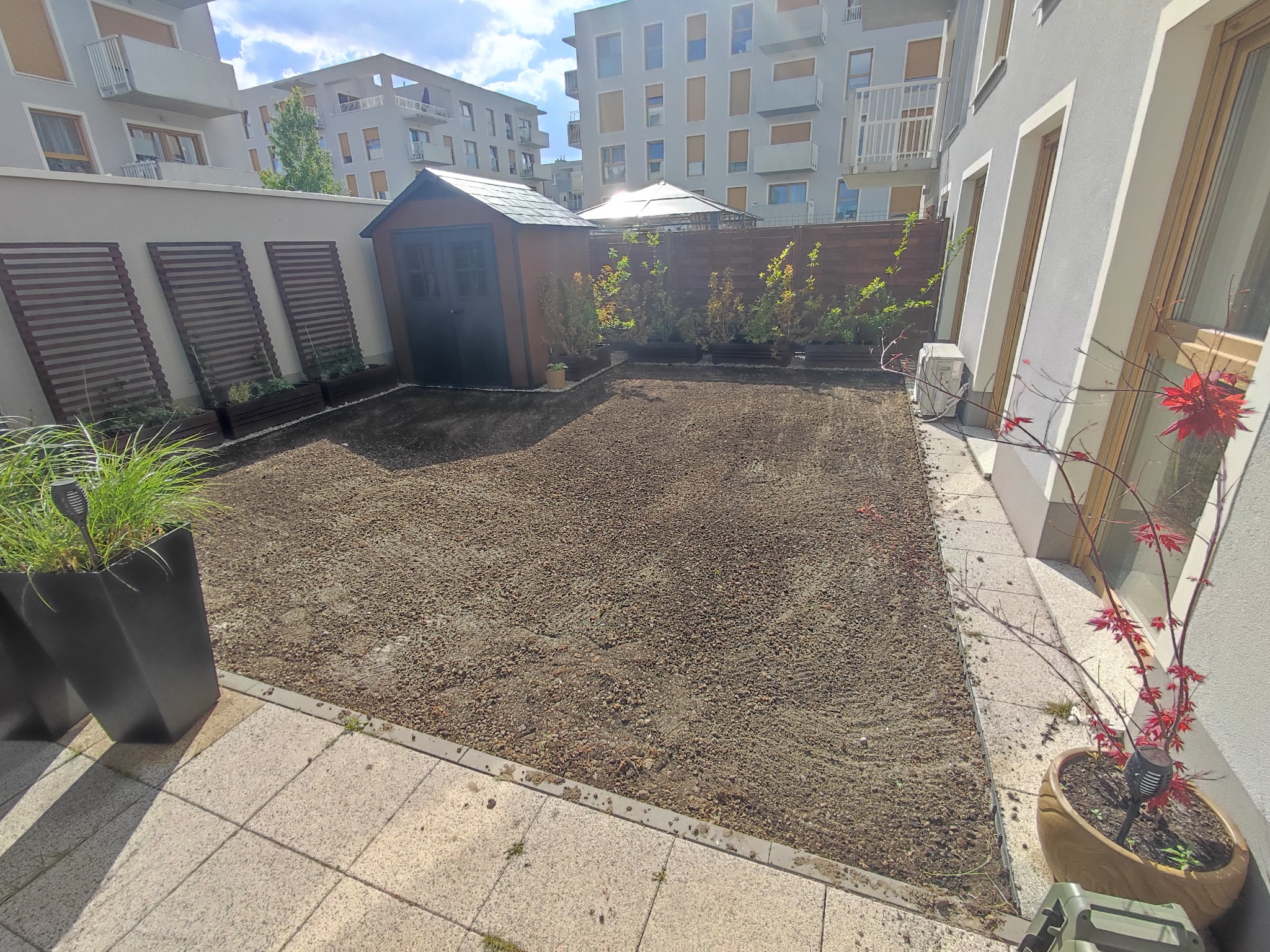 Przygotowany teren pod trawnik w Kostomłotach, widoczne wyrównane podłoże, obrzeża z kostki, donice z roślinami i budynek gospodarczy w tle.