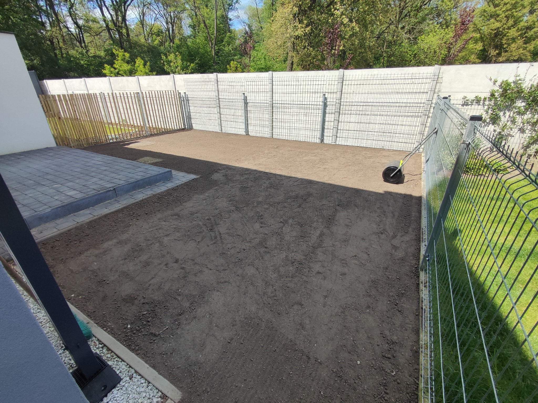 Przygotowany teren pod trawnik, wyrównana ziemia, widoczny walec ogrodowy, ogrodzenie panelowe w tle, częściowo ułożona kostka brukowa.