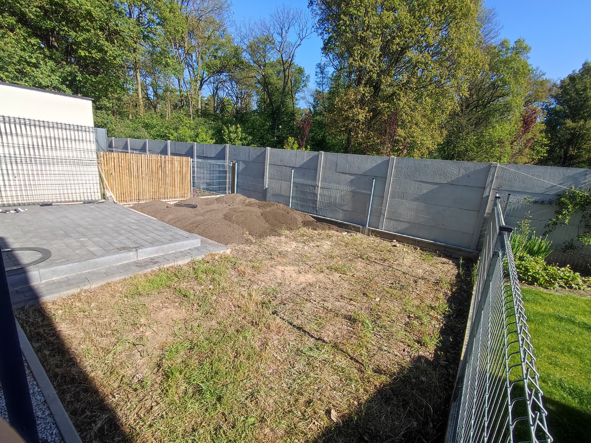 Przygotowanie terenu pod trawnik: wyrównana ziemia, widoczna kostka brukowa, ogrodzenie panelowe i betonowe w tle z zielenią.