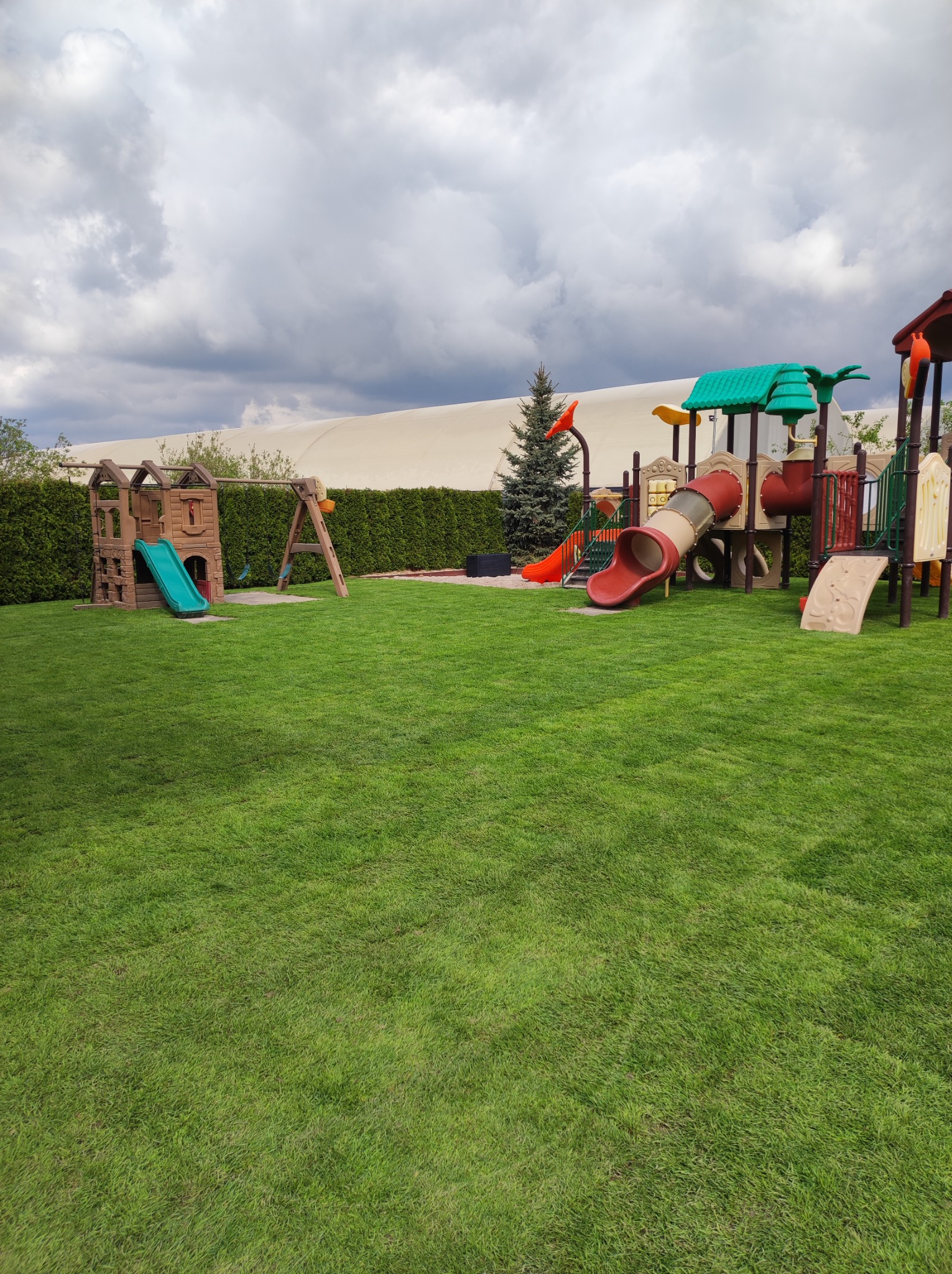 Rozległy, zielony trawnik z równo skoszoną trawą, otoczony żywopłotem i wyposażony w kolorowy plac zabaw z drewnianą konstrukcją, zjeżdżalniami i huśtawkami, na tle pochmurnego nieba.