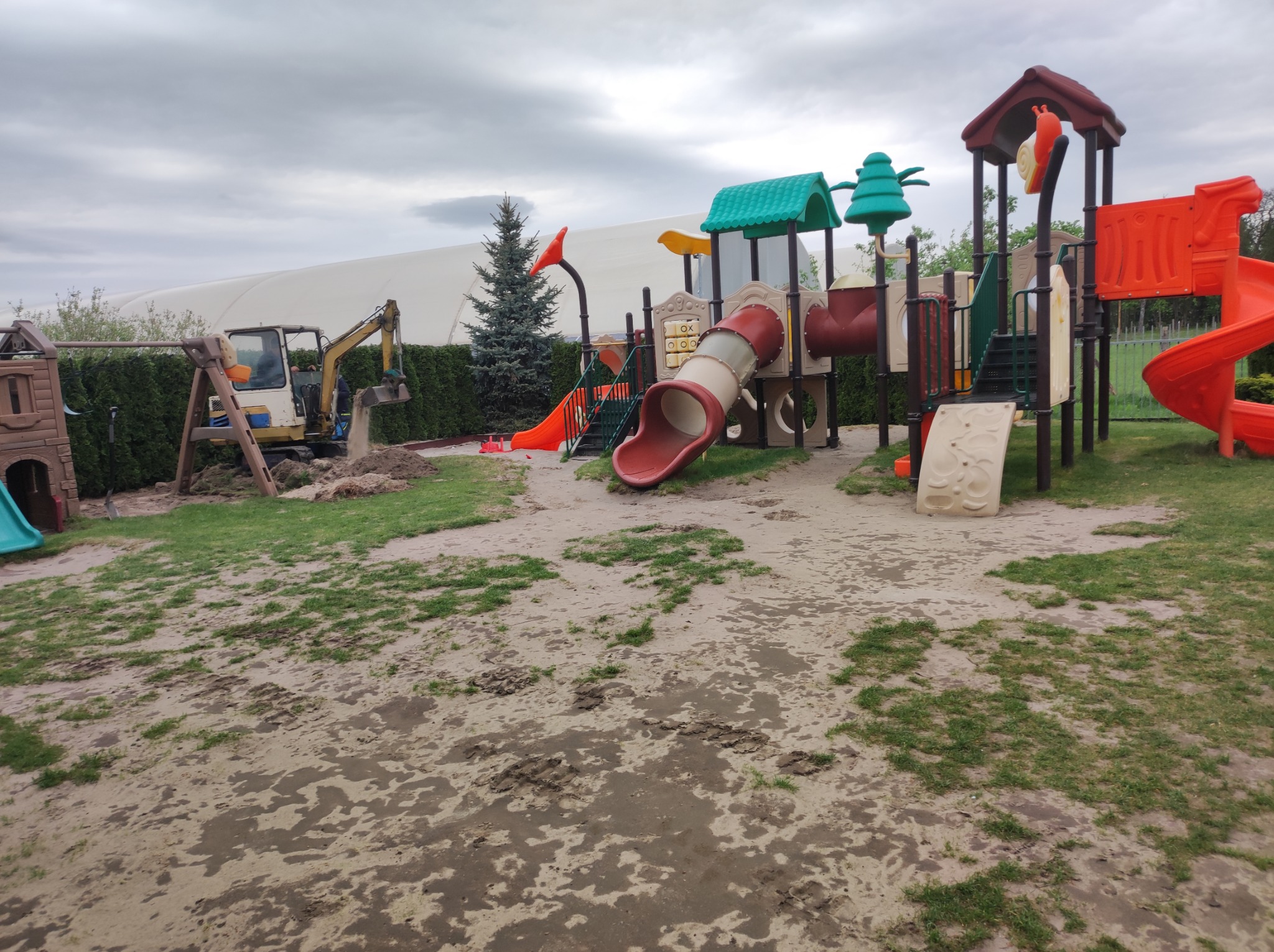 Żółta koparka pracuje na placu zabaw, przygotowując teren pod instalację urządzeń, w tle widoczny duży namiot.