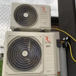 Dwie jednostki zewnętrzne klimatyzacji Rotenso, model Unico i Hiro, zamontowane jedna nad drugą, obok butla z gazem i manometry, widoczne przewody, tło stanowi szara ściana i siatka ogrodzeniowa.