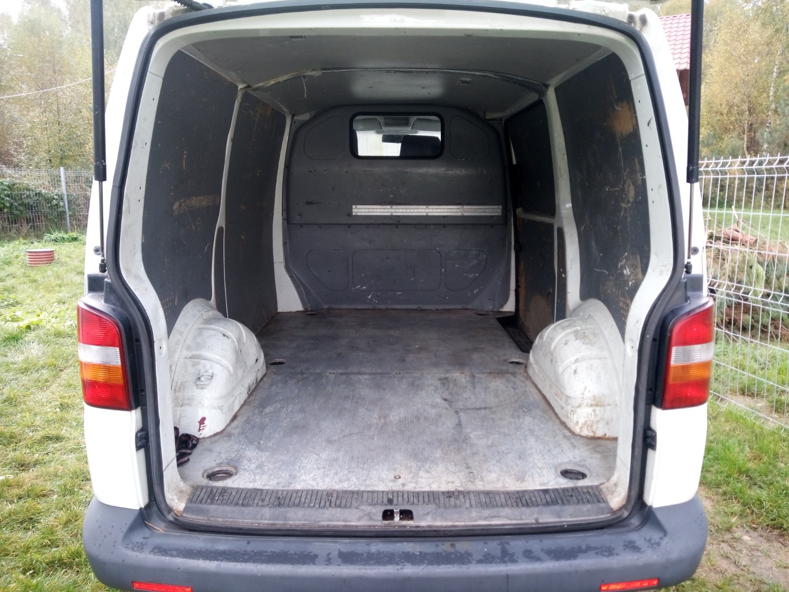 Wnętrze puste, brudne przestrzeni ładunkowej białego busa z otwartymi drzwiami, widok z tyłu, z elementami mocującymi w podłodze i śladami użytkowania na ścianach.