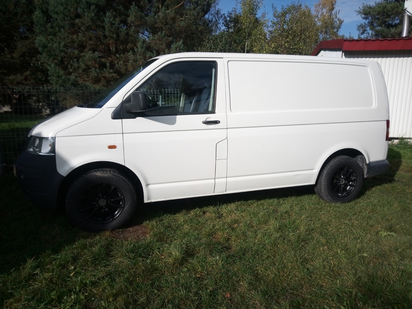 Biały samochód dostawczy Volkswagen Transporter T5 z czarnymi felgami, zaparkowany na trawie przed budynkiem z czerwonym dachem.