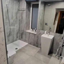 Nowoczesna łazienka z szarymi płytkami imitującymi kamień, kabiną prysznicową typu walk-in, wiszącą umywalką o geometrycznym kształcie i lustrem powiększającym przestrzeń.