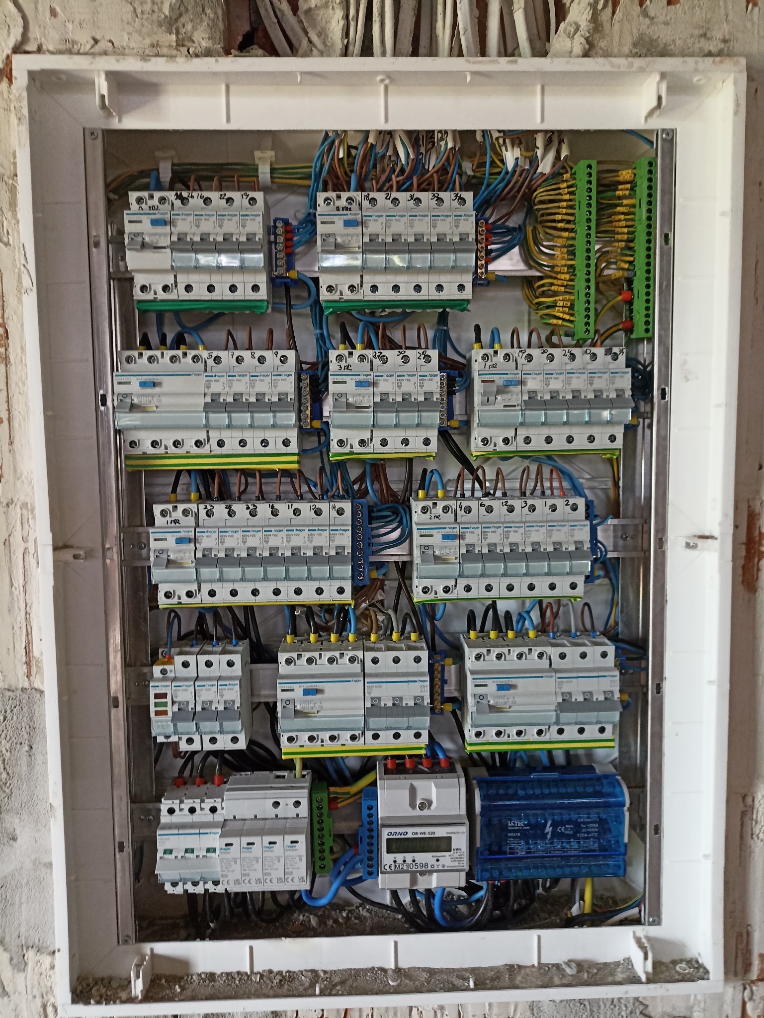 Rozdzielnica elektryczna Hager z zamontowanymi bezpiecznikami, przewodami i licznikiem ORNO OR-WE-520, widok z bliska na tle surowej ściany w trakcie remontu.