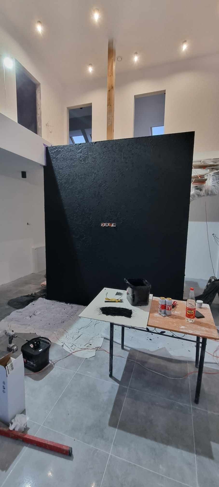 Czarna, teksturowana ściana wewnętrzna w trakcie malowania z widocznymi gniazdkami elektrycznymi, narzędzia malarskie i puszki z napojami na prowizorycznym stole, podłoga wyłożona szarymi płytkami.
