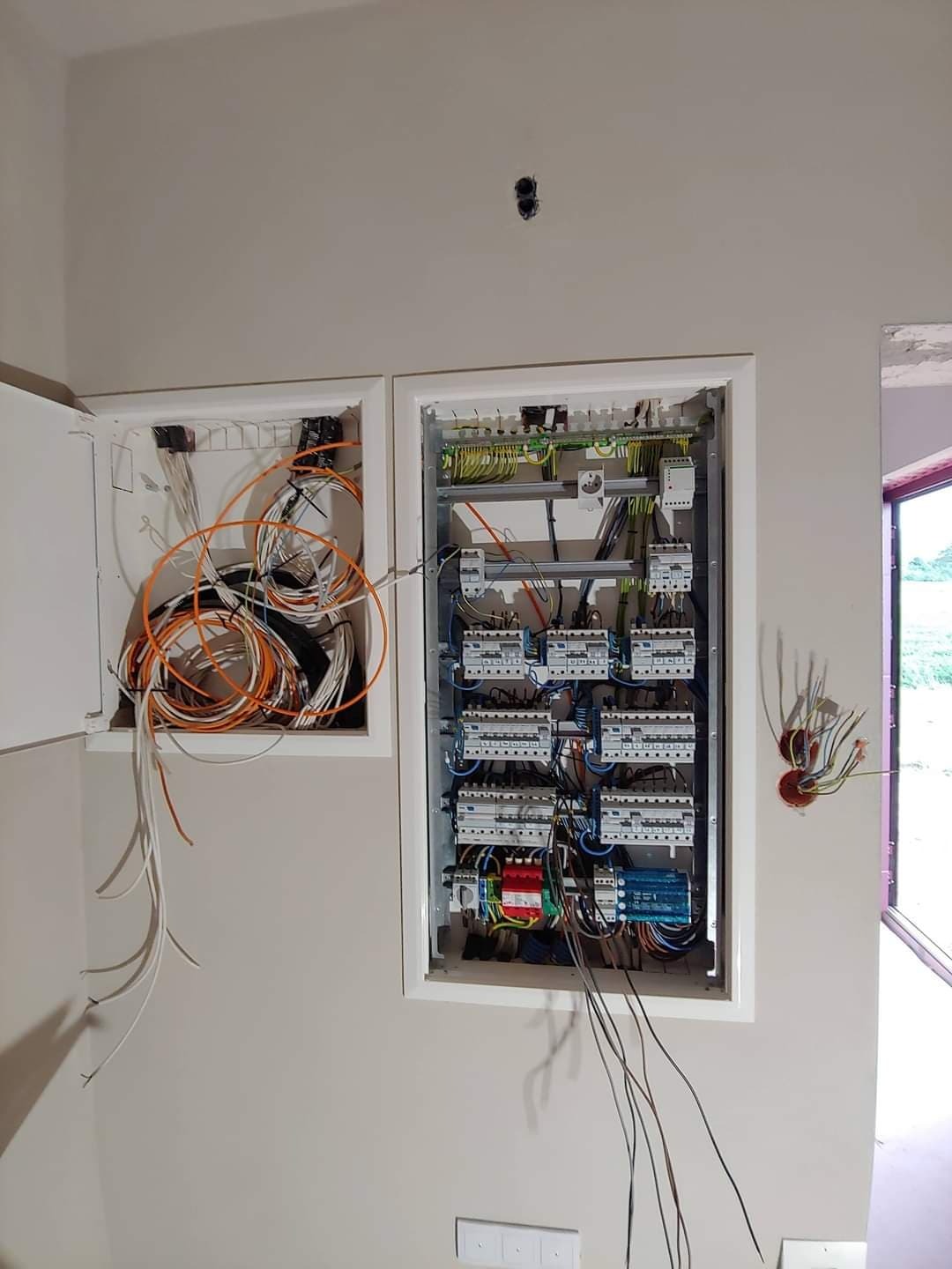 Rozdzielnica elektryczna z zabezpieczeniami i okablowaniem w dwóch wnękach ściennych, widoczne przewody w różnych kolorach, część obudowy otwarte, jasne ściany w tle.