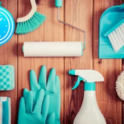 Zestaw narzędzi do sprzątania w odcieniach błękitu i bieli, ułożony na drewnianym tle: szczotka, zmiotka, wałek do malowania, spryskiwacz, gąbka, ściereczka i rękawice gumowe.