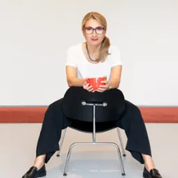 Kobieta w okularach siedzi na czarnym krześle, trzymając czerwoną filiżankę, ubrana w białą bluzkę i czarne spodnie.