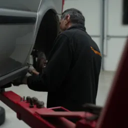 Szary van na podnośniku samochodowym, pracownik w czarnej bluzie z napisem 'TANSERVICE' na plecach, w rękawicach pracuje przy zawieszeniu.