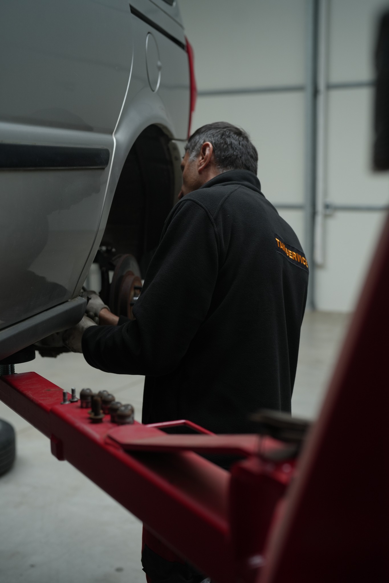 Szary van na podnośniku samochodowym, pracownik w czarnej bluzie z napisem 'TANSERVICE' na plecach, w rękawicach pracuje przy zawieszeniu.