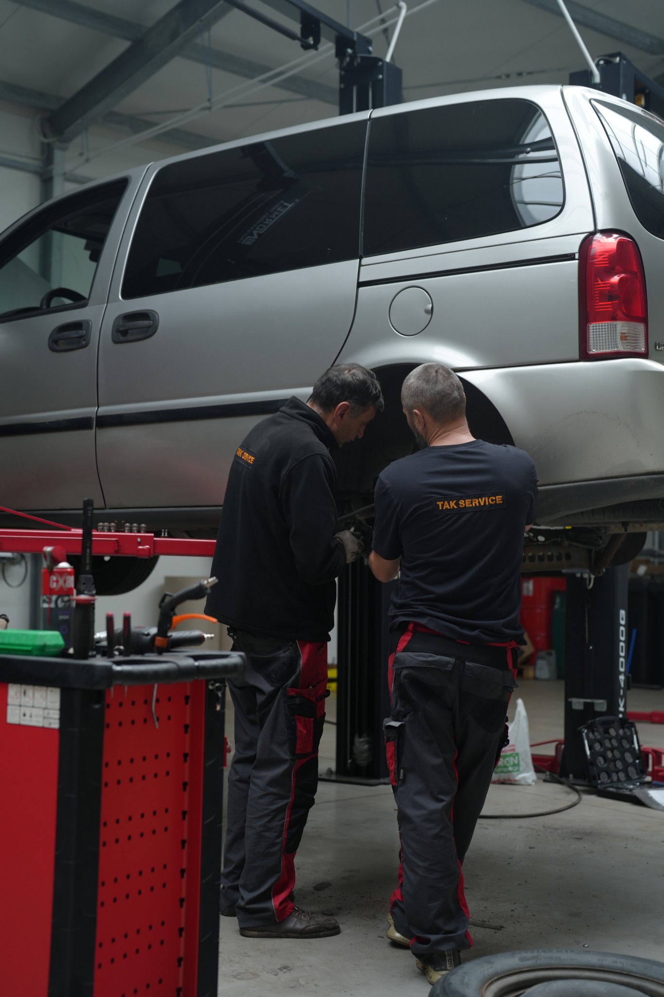 Dwóch pracowników warsztatu samochodowego w firmowych strojach, pracujących przy podniesionym srebrnym minivanie na podnośniku, widoczny wózek narzędziowy i opona na podłodze.