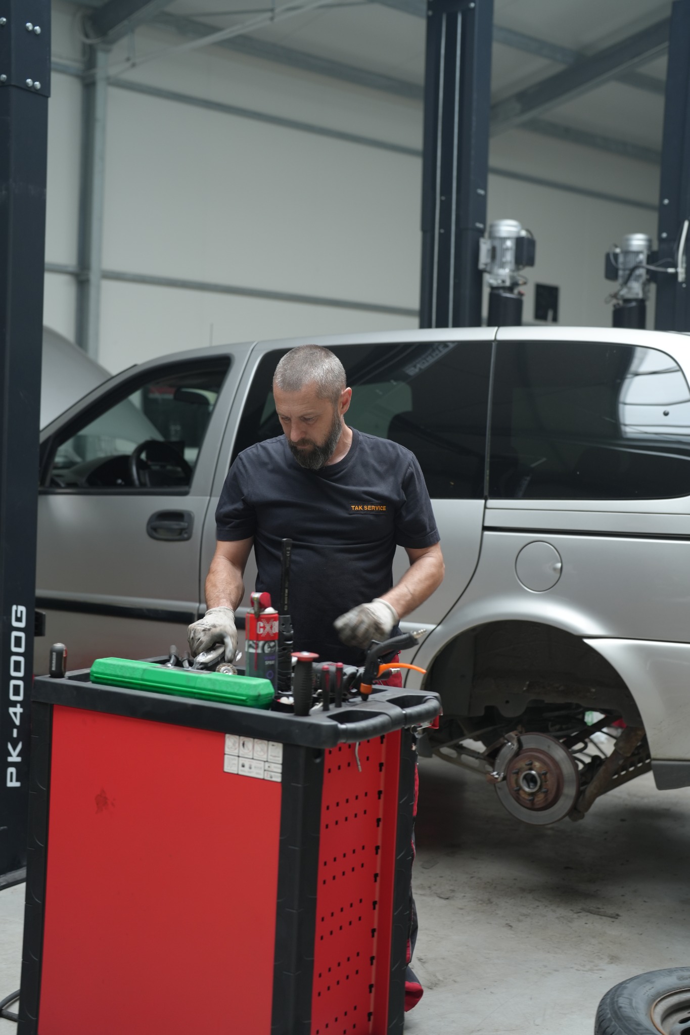 Mężczyzna w warsztacie samochodowym, ubrany w roboczą koszulkę z napisem 'TAK SERVICE', pochyla się nad wózkiem narzędziowym z zielonym pojemnikiem, obok podnośnika i srebrnego samochodu bez koła...