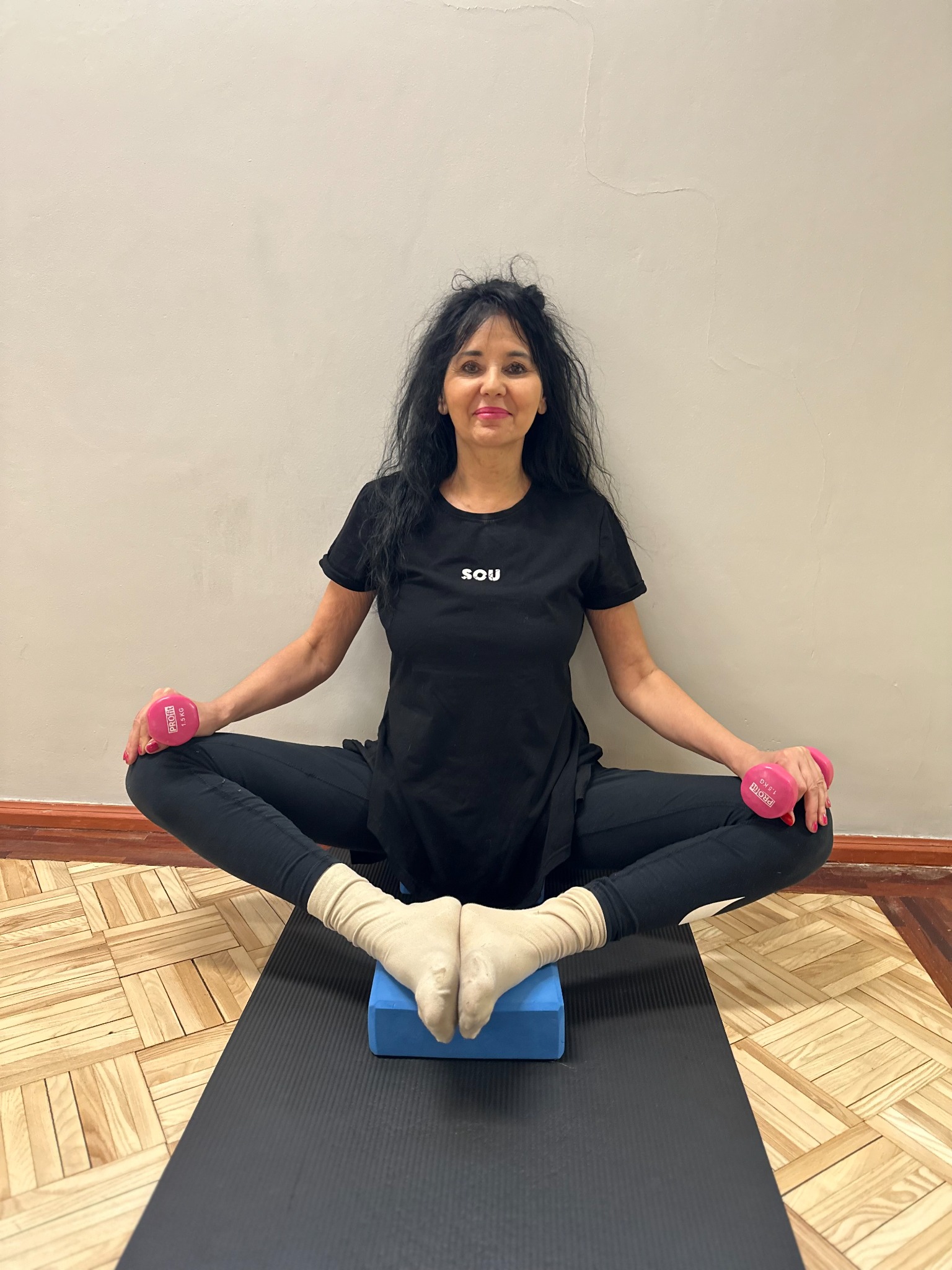 Kobieta w czarnym stroju siedzi na macie do ćwiczeń, trzymając w dłoniach różowe hantle, stopy oparte o niebieski blok do jogi, na drewnianej podłodze.